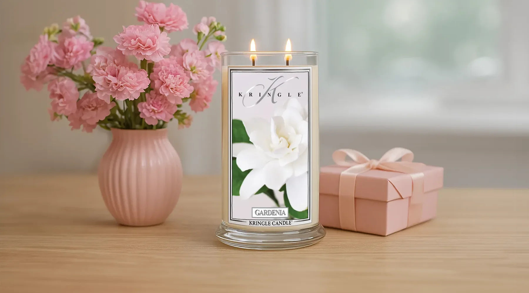 Kringle Candle Gardenia candle with two flames lit, placed on a wooden table beside a pink vase of carnations and a pink ribbon-wrapped gift box in a softly lit room.