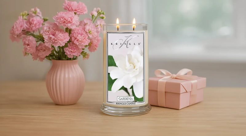 Kringle Candle Gardenia candle with two flames lit, placed on a wooden table beside a pink vase of carnations and a pink ribbon-wrapped gift box in a softly lit room.