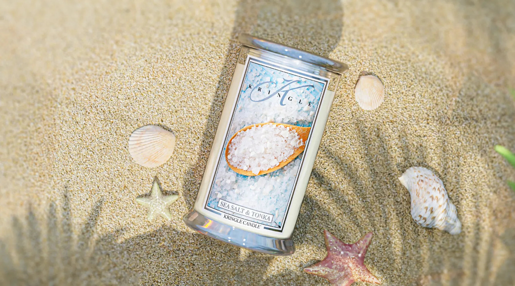 Kringle Candle’s Sea Salt & Tonka large jar candle resting on sunlit sand, surrounded by seashells.