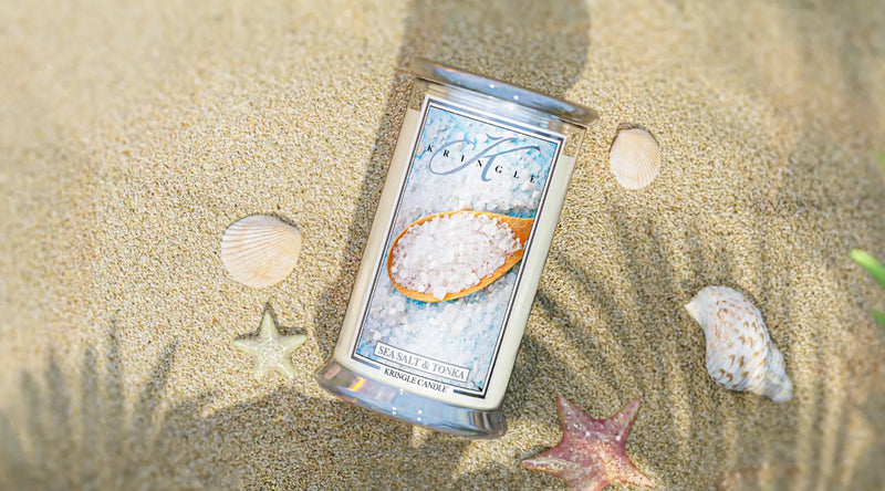 Kringle Candle’s Sea Salt & Tonka large jar candle resting on sunlit sand, surrounded by seashells.