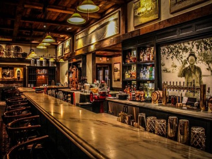 Interior of the bar at The Farm Table Restaurant featuring a long bar with high-backed chairs, warm ambient lighting, shelves stocked with liquor bottles, multiple beer taps, and rustic décor including vintage photographs and wooden beams.