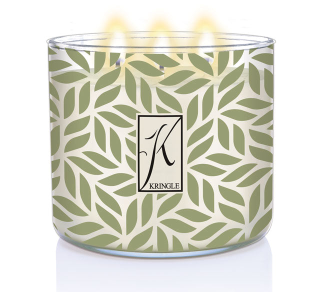 A lit candle in a glass container with a green leaf pattern and a stylized "K" logo in the center.