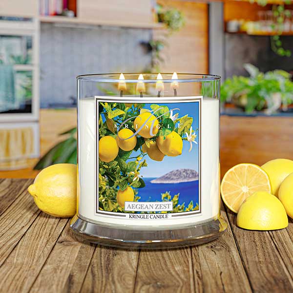 Kringle Candle Aegean Zest 4-Wick Jar Candle – A wide, clear glass jar candle with four lit wicks, filled with white wax. The label showcases vibrant yellow lemons, green leaves, and white blossoms against a deep blue ocean and an island in the background. The ‘Kringle Candle’ logo is displayed at the top, with the fragrance name ‘Aegean Zest’ in bold, and ‘Kringle Candle’ branding at the bottom.
