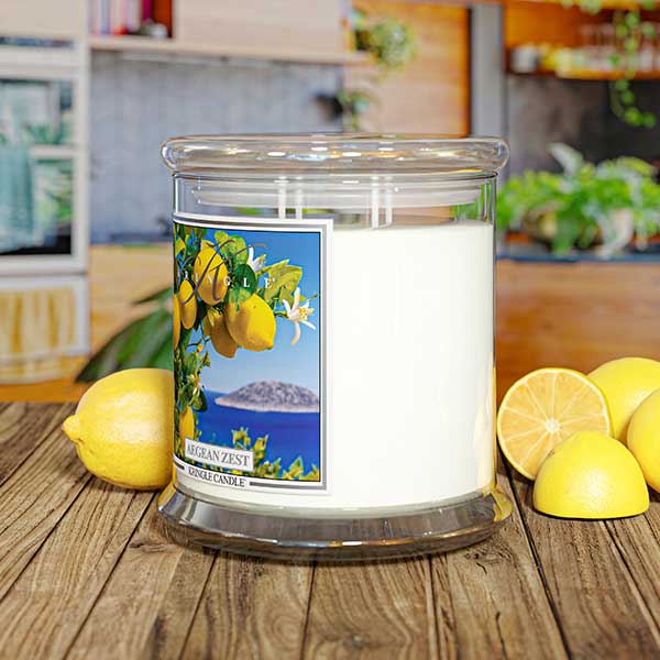 Kringle Candle Aegean Zest 4-Wick Jar Candle – A wide, clear glass jar candle with four lit wicks, filled with white wax. The label showcases vibrant yellow lemons, green leaves, and white blossoms against a deep blue ocean and an island in the background. The ‘Kringle Candle’ logo is displayed at the top, with the fragrance name ‘Aegean Zest’ in bold, and ‘Kringle Candle’ branding at the bottom.