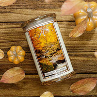 Kringle Candle with 'Amberwood Moss' candle on a wooden surface with a blurred autumn-themed background.