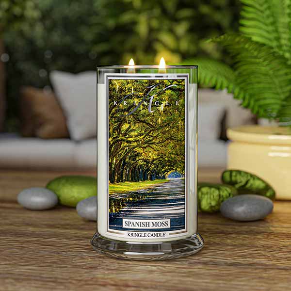 A lit Kringle Candle jar labeled “Spanish Moss” placed on a wooden surface surrounded by smooth gray and green stones, a yellow ceramic pot, and lush green ferns. The candle’s label displays a serene tree-lined path covered in Spanish moss. A cozy, blurred background with cushions enhances the peaceful, nature-inspired indoor setting.