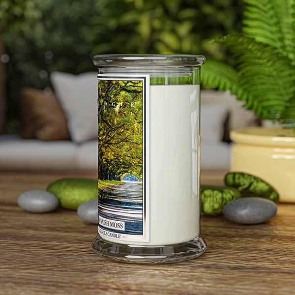 A Kringle Candle jar labeled “Spanish Moss” standing upright on a wooden surface. The candle is surrounded by smooth gray and green stones, a potted plant, and fern leaves. The background features a cozy, blurred-out indoor seating area, evoking a calm and natural ambiance.