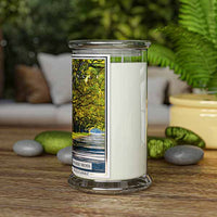 A Kringle Candle jar labeled “Spanish Moss” standing upright on a wooden surface. The candle is surrounded by smooth gray and green stones, a potted plant, and fern leaves. The background features a cozy, blurred-out indoor seating area, evoking a calm and natural ambiance.