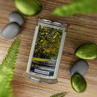 A Kringle Candle jar labeled “Spanish Moss” placed on a wooden surface, surrounded by smooth gray and mossy green stones and fern leaves. The candle label features a scenic path lined with large, arching oak trees draped in Spanish moss, evoking a peaceful, natural Southern landscape.