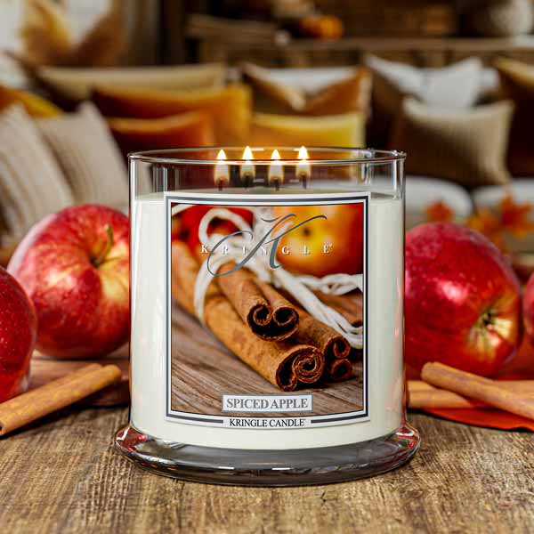 Kringle Candle with scented 4-wick'Spiced Apple' label surrounded by apples and cinnamon sticks on a wooden surface