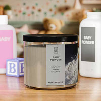 A three-wick candle in a clear glass jar with a black lid and a classic powder-themed label. The label features a bottle of baby powder spilling onto a dark surface. The candle’s wax is soft white, and the wicks are unlit. The scent notes listed are baby powder, lavender, citrus, and floral.