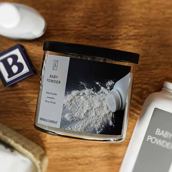 A three-wick candle in a clear glass jar with a black lid and a classic powder-themed label. The label features a bottle of baby powder spilling onto a dark surface. The candle’s wax is soft white, and the wicks are unlit. The scent notes listed are baby powder, lavender, citrus, and floral.