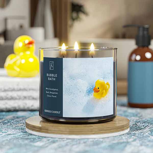 A three-wick lit candle in a clear glass jar with a calming bath-themed label. The candle’s wax is dark blue, and all three wicks are burning.