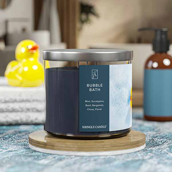 A three-wick candle in a clear glass jar with a silver lid and a calming bath-themed label. The label features a rubber duck floating in a sea of white bubbles. The candle’s wax is dark blue, and the wicks are unlit. The scent notes listed are mint, eucalyptus, basil, bergamot, clove, and floral.