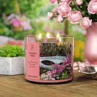 A three-wick lit candle in a clear glass jar with a serene garden-themed label. The candle’s wax is a soft pink shade, and all three wicks are burning.