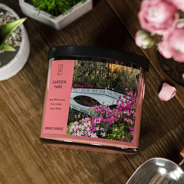 A three-wick candle in a clear glass jar with a black lid and a serene garden-themed label. The label features a white bridge arching over a pond, surrounded by blooming pink flowers and lush greenery. The candle’s wax is a soft pink shade, and the wicks are unlit. The scent notes listed are rose, white lotus, floral, violet, moss, and wood.