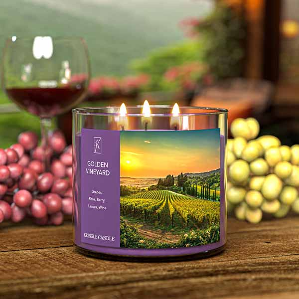 A three-wick lit candle in a clear glass jar with a scenic vineyard-themed label. The candle’s wax is deep purple, and all three wicks are burning.