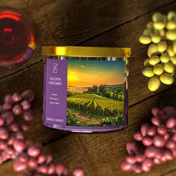 A three-wick candle in a clear glass jar with a gold lid and a scenic vineyard-themed label. The label features a picturesque vineyard at sunset with rolling green hills and a golden sky. The candle’s wax is deep purple, and the wicks are unlit. The scent notes listed are grapes, rose, berry, leaves, and wine.