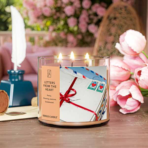 A three-wick lit candle in a clear glass jar with a sentimental-themed label. The candle’s wax is a soft peach shade, and all three wicks are burning.