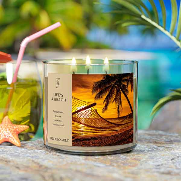 A three-wick lit candle in a clear glass jar with a tropical sunset-themed label. The candle’s wax is sandy beige, and all three wicks are burning.