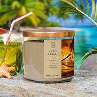 A three-wick candle in a clear glass jar with a bronze lid and a tropical sunset-themed label. The label features a hammock tied between palm trees on a golden beach at sunset, with warm orange hues in the sky. The candle’s wax is sandy beige, and the wicks are unlit. The scent notes listed are tonka bean, amber, jasmine, and sandalwood.