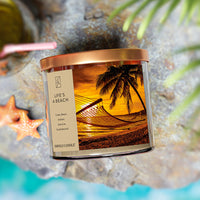 A three-wick candle in a clear glass jar with a bronze lid and a tropical sunset-themed label. The label features a hammock tied between palm trees on a golden beach at sunset, with warm orange hues in the sky. The candle’s wax is sandy beige, and the wicks are unlit. The scent notes listed are tonka bean, amber, jasmine, and sandalwood.