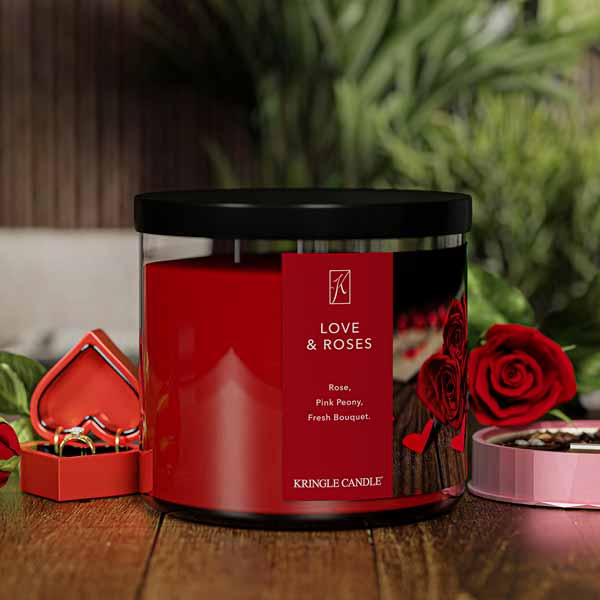 A three-wick lit candle in a clear glass jar with a romantic floral-themed label. The candle’s wax is deep red, and the jar has a black lid.