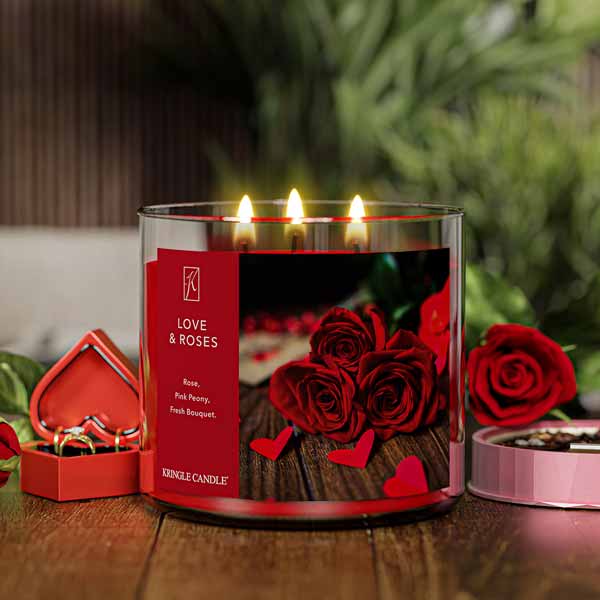 A three-wick lit candle in a clear glass jar with a romantic floral-themed label. The candle’s wax is deep red, and all three wicks are burning.
