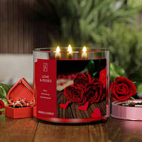 A three-wick lit candle in a clear glass jar with a romantic floral-themed label. The candle’s wax is deep red, and all three wicks are burning.