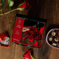 A three-wick lit candle in a clear glass jar with a romantic floral-themed label. The candle’s wax is deep red, and the jar has a black lid.
