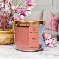 A three-wick candle in a clear glass jar with a bronze lid and a soft floral-themed label. The label features a close-up of pink magnolia blossoms against a bright blue sky. The candle’s wax is a warm peach shade, and the wicks are unlit. The scent notes listed are magnolia, fresh leaves, and lemon.