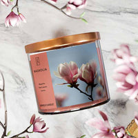 A three-wick candle in a clear glass jar with a bronze lid and a soft floral-themed label. The label features a close-up of pink magnolia blossoms against a bright blue sky. The candle’s wax is a warm peach shade, and the wicks are unlit. The scent notes listed are magnolia, fresh leaves, and lemon.