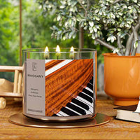 A three-wick lit candle in a clear glass jar with a luxurious wood and music-themed label. The candle’s wax is a rich brown, and all three wicks are burning.