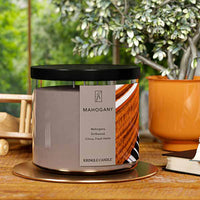 A three-wick candle in a clear glass jar with a black lid and a luxurious wood and music-themed label. The label features a polished mahogany piano with black and white keys, showcasing the deep reddish-brown wood grain. The candle’s wax is a rich brown, and the wicks are unlit. The scent notes listed are mahogany, driftwood, citrus, and fresh herbs.