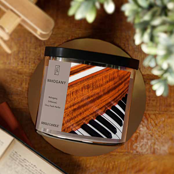 A three-wick candle in a clear glass jar with a black lid and a luxurious wood and music-themed label. The label features a polished mahogany piano with black and white keys, showcasing the deep reddish-brown wood grain. The candle’s wax is a rich brown, and the wicks are unlit. The scent notes listed are mahogany, driftwood, citrus, and fresh herbs.