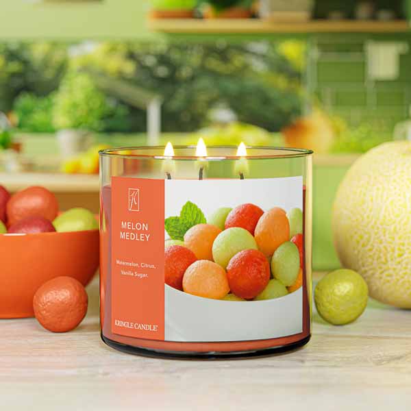 A three-wick lit candle in a clear glass jar with a fresh fruit-themed label. The candle’s wax is warm orange, and all three wicks are burning.