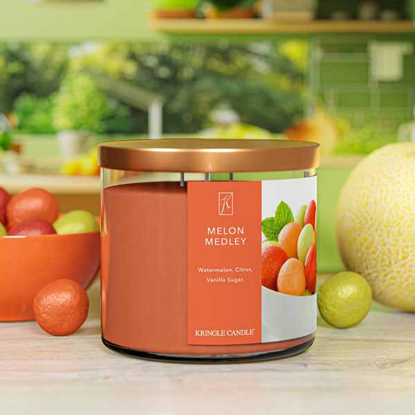 A three-wick candle in a clear glass jar with a bronze lid and a fresh fruit-themed label. The label features a bowl filled with colorful melon balls, including watermelon, cantaloupe, and honeydew. The candle’s wax is warm orange, and the wicks are unlit. The scent notes listed are watermelon, citrus, and vanilla sugar.