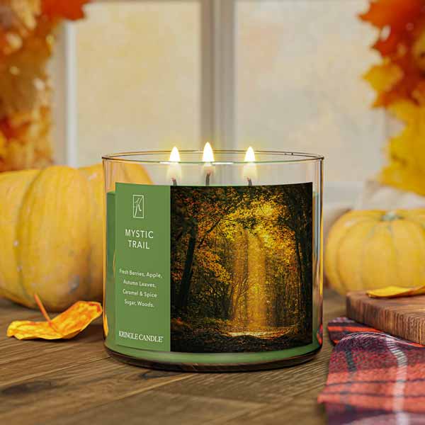 A three-wick lit candle in a clear glass jar with a forest-inspired label. The candle’s wax is an earthy green, and all three wicks are burning.