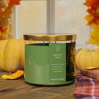 A three-wick candle in a clear glass jar with a gold lid and a forest-inspired label. The label features a golden-lit forest path with autumn leaves and sun rays filtering through the trees. The candle’s wax is an earthy green, and the wicks are unlit. The scent notes listed are fresh berries, apple, autumn leaves, caramel & spice sugar, and woods.