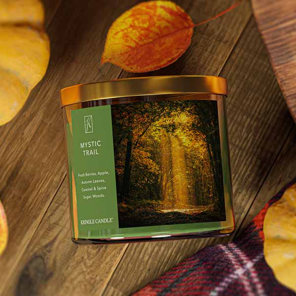 A three-wick candle in a clear glass jar with a gold lid and a forest-inspired label. The label features a golden-lit forest path with autumn leaves and sun rays filtering through the trees. The candle’s wax is an earthy green, and the wicks are unlit. The scent notes listed are fresh berries, apple, autumn leaves, caramel & spice sugar, and woods.