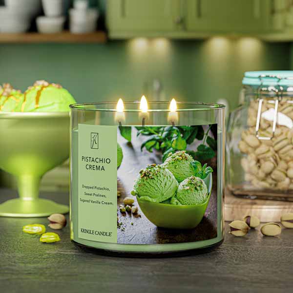A three-wick lit candle in a clear glass jar with a creamy dessert-themed label. The candle’s wax is soft green, and all three wicks are burning.