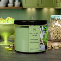 A three-wick candle in a clear glass jar with a gold lid and a creamy dessert-themed label. The label features a bowl of pistachio gelato topped with chopped nuts and a swirl of caramel. The candle’s wax is soft green, and the wicks are unlit. The scent notes listed are pistachio, pralines, and sugared vanilla cream.