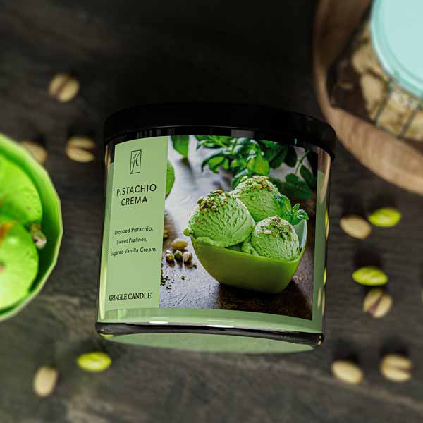 A three-wick candle in a clear glass jar with a gold lid and a creamy dessert-themed label. The label features a bowl of pistachio gelato topped with chopped nuts and a swirl of caramel. The candle’s wax is soft green, and the wicks are unlit. The scent notes listed are pistachio, pralines, and sugared vanilla cream.