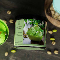 A three-wick candle in a clear glass jar with a gold lid and a creamy dessert-themed label. The label features a bowl of pistachio gelato topped with chopped nuts and a swirl of caramel. The candle’s wax is soft green, and the wicks are unlit. The scent notes listed are pistachio, pralines, and sugared vanilla cream.
