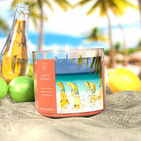 A three-wick lit candle in a clear glass jar with a tropical beach-themed label. The candle’s wax is soft coral, and all three wicks are burning.