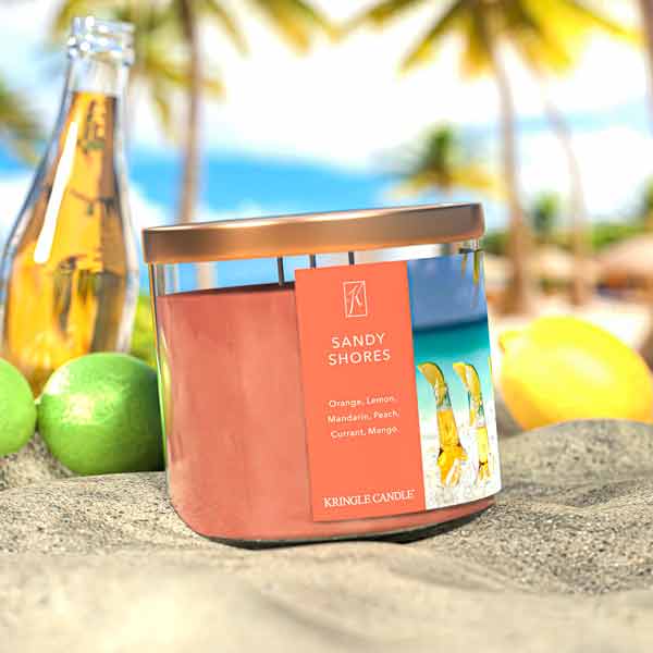 A three-wick candle in a clear glass jar with a bronze lid and a tropical beach-themed label. The label features a serene shoreline with golden sand, turquoise water, and a bright blue sky. The candle’s wax is soft coral, and the wicks are unlit. The scent notes listed are orange, lemon, peach, mandarin, mango, and red currant.