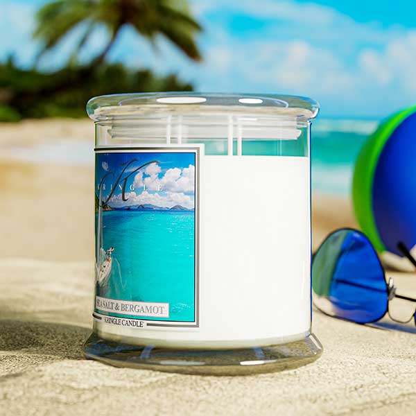 A Kringle Candle jar labeled “Sea Salt & Bergamot” placed on sandy ground at the beach. The background features a turquoise ocean, palm trees, and blue sky. A pair of sunglasses and a colorful beach ball are positioned beside the candle, evoking a relaxed, tropical atmosphere.