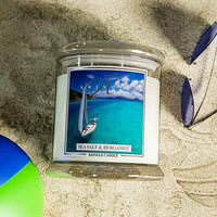 A Kringle Candle jar labeled “Sea Salt & Bergamot” placed on sandy ground with a beach ball partially visible in the lower left and blue leaves in the upper right. The candle label features a sailboat gliding over turquoise water with an island and blue sky in the background, evoking a tropical, oceanic vibe.