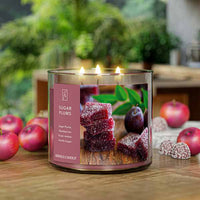 A three-wick lit candle in a clear glass jar with a festive holiday-themed label. The candle’s wax is deep purple, and all three wicks are burning.