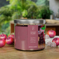 A three-wick candle in a clear glass jar with a silver lid and a festive holiday-themed label. The label features a bowl of sugared plums and juicy blackberries, dusted with powdered sugar. The candle’s wax is deep purple, and the wicks are unlit. The scent notes listed are sugared plums, blackberries, delicate florals, amber, and vanilla sugar.